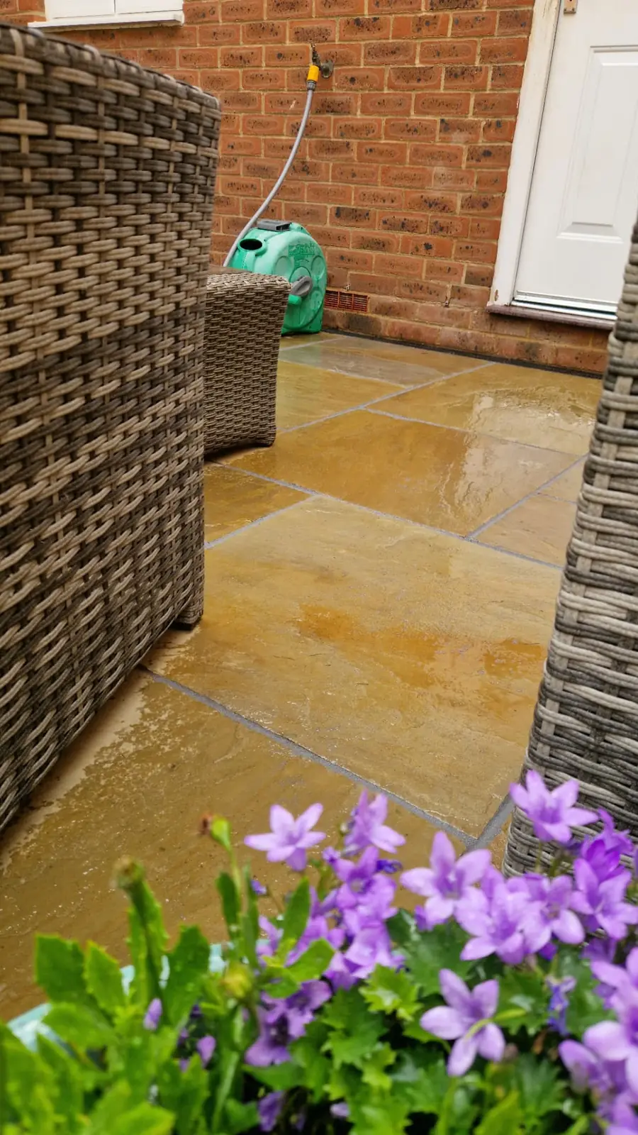 Freshly cleaned Indian sandstone patio, wet after washing, with rattan furniture and a pot of campanula